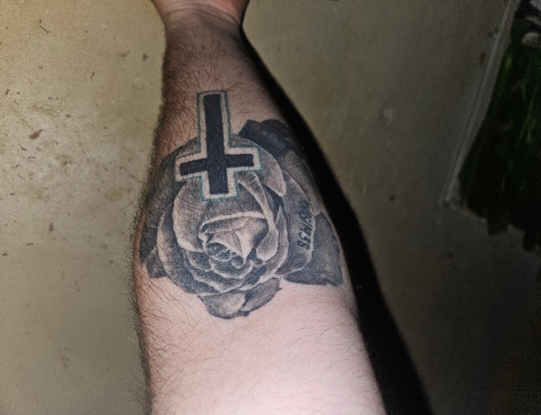 A tattoo on an arm featuring a black rose and an inverted cross. The date '06/1958' is visible on the rose.