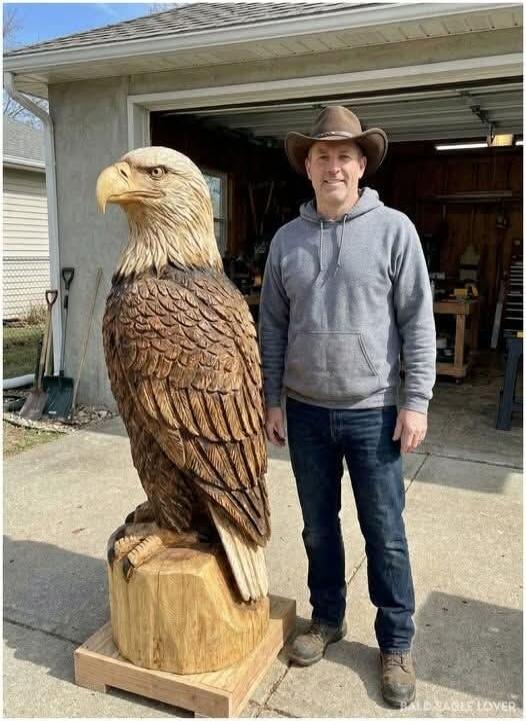 A man in a cowboy hat and a grey hoodie stands next to a large, impressive wooden sculpture of a bald eagle.