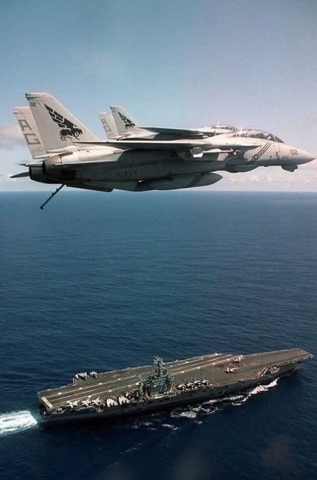 Fighter jet flying above an aircraft carrier at sea.