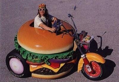 A man driving a custom vehicle shaped like a giant hamburger. The vehicle features a motorcycle front end and a large burger body with ingredients like lettuce, cheese, and tomato. The rear tires have 'Radial T/A' and 'Goodyear' visible.