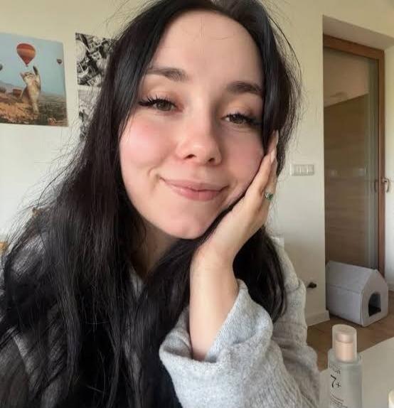 A young woman with long dark hair smiles at the camera, resting her chin on her hand. She is wearing a grey knitted sweater. In the background, there are framed pictures, including one of a cat in a hot air balloon, and a pet bed. A bottle of '7+' skincare product is visible on a table.