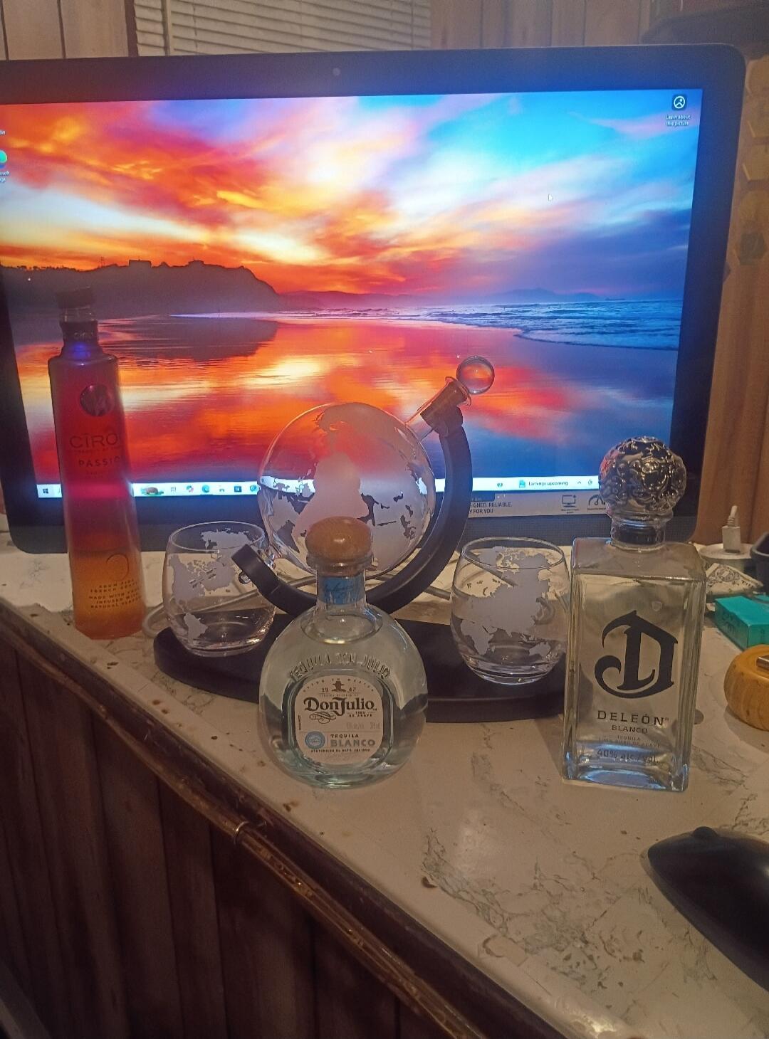 Ciroc Passion, Don Julio Tequila Blanco, DeLeon Blanco Tequila. A collection of spirits displayed on a desk with a globe decanter and glasses, in front of a computer monitor showing a sunset beach wallpaper.