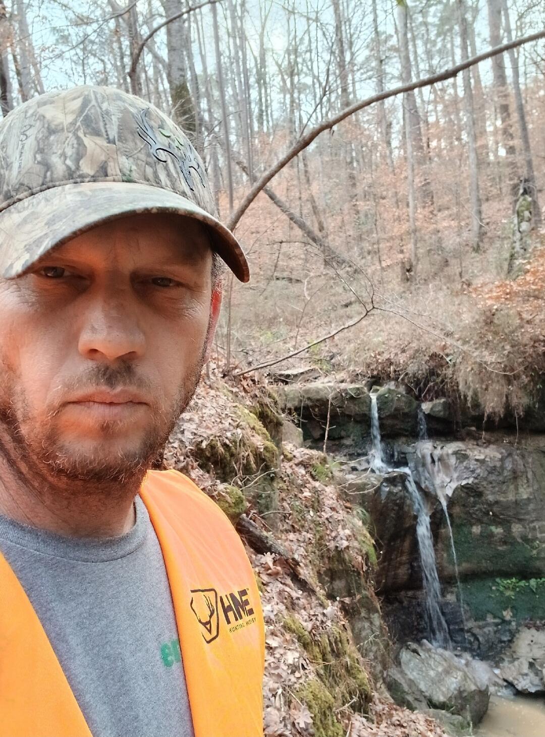 A man in a camouflage hat and an orange vest with 'HHE HUNTING HOBBY' text, standing in a forest with a small waterfall in the background.