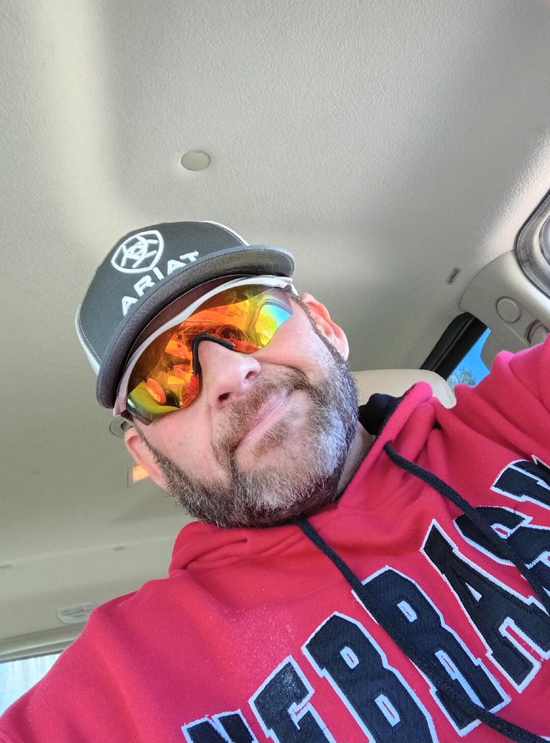 A man wearing an Ariat cap, mirrored sunglasses, and a red Nebraska hoodie takes a selfie in a car.