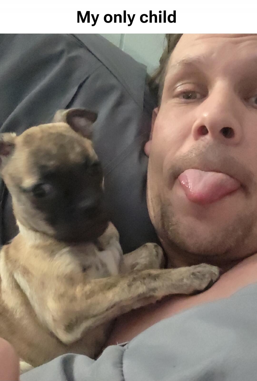 A man takes a selfie with a small puppy resting on his chest. The man is playfully sticking out his tongue. The text overlay reads: 
