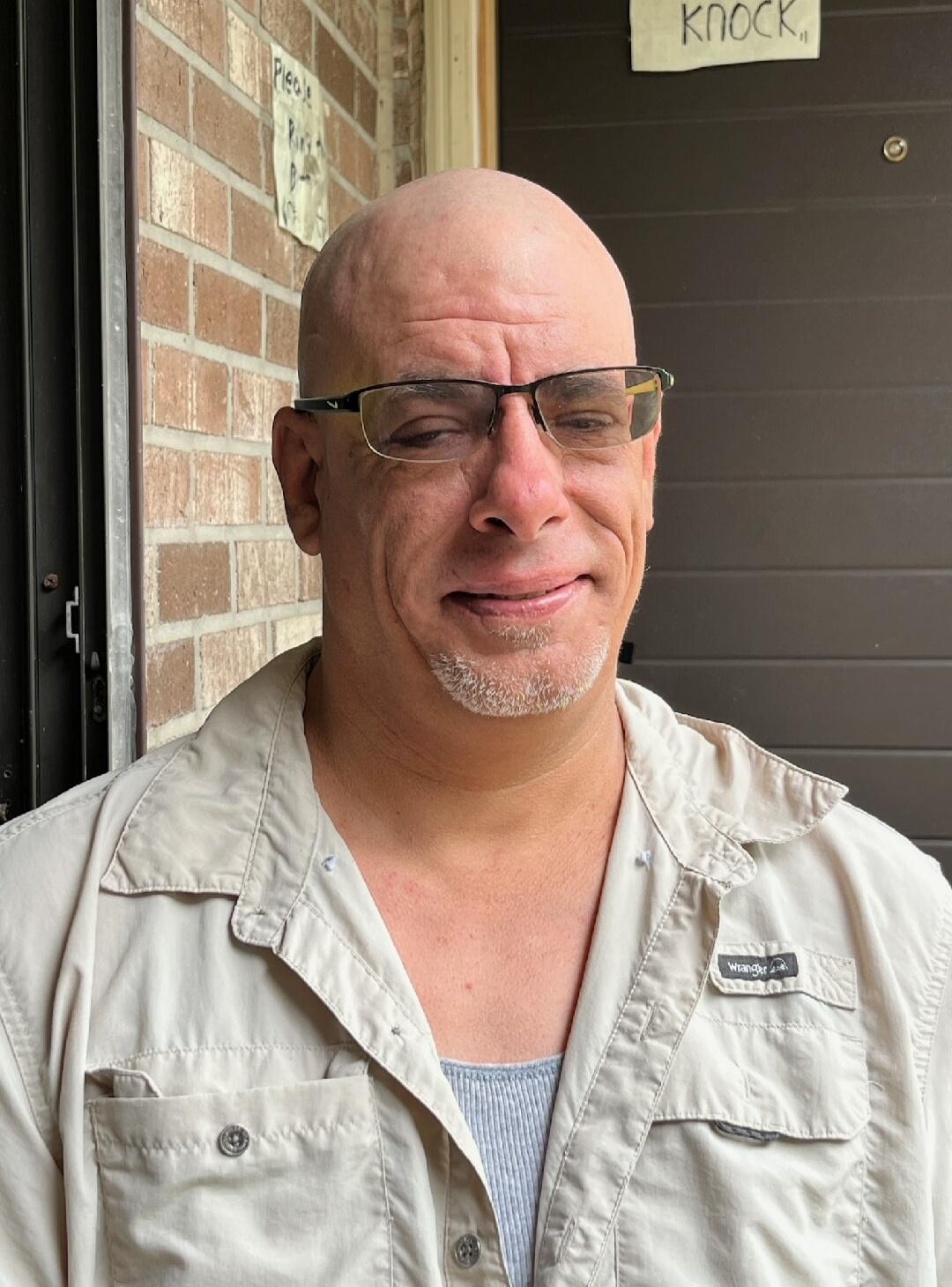 Please Ring, KNOCK. A bald man with glasses and a goatee stands in front of a brick wall and a door, with a slight smirk on his face.