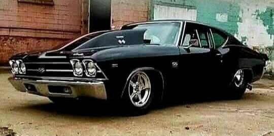 A black classic muscle car, possibly a Chevrolet Chevelle, with a large hood scoop and custom wheels, parked in front of a building.