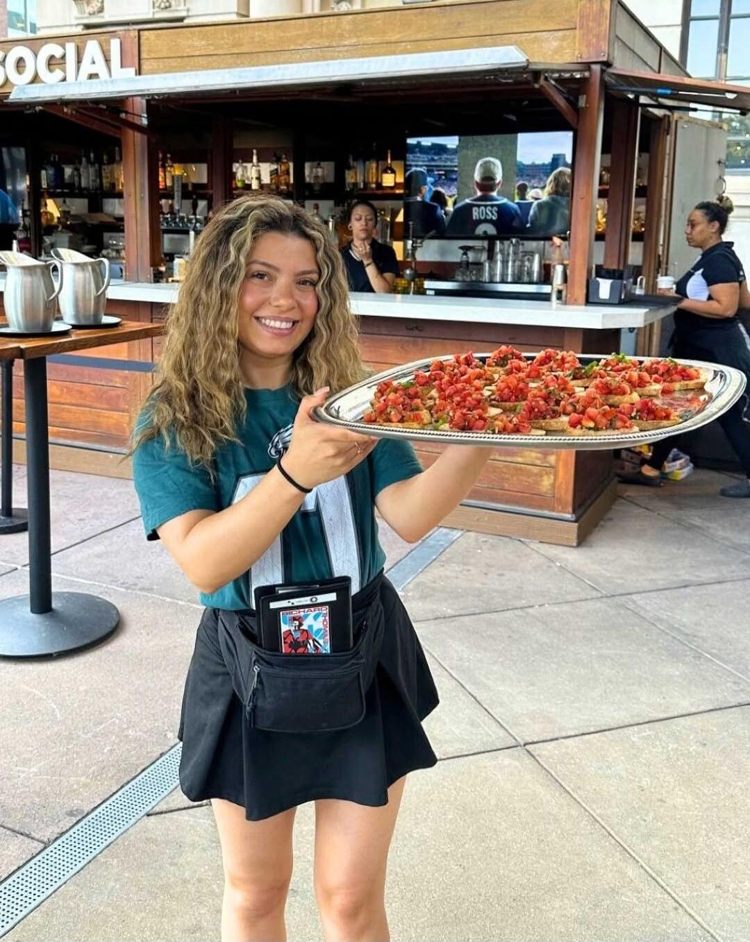 A smiling woman, likely a server, holds a large silver tray filled with numerous servings of bruschetta. She is wearing a green t-shirt, a black pleated skirt, and a fanny pack containing a tablet or POS device. The background features an outdoor bar area with the sign 'SOCIAL' visible, and a television screen displaying a sports game where a playe