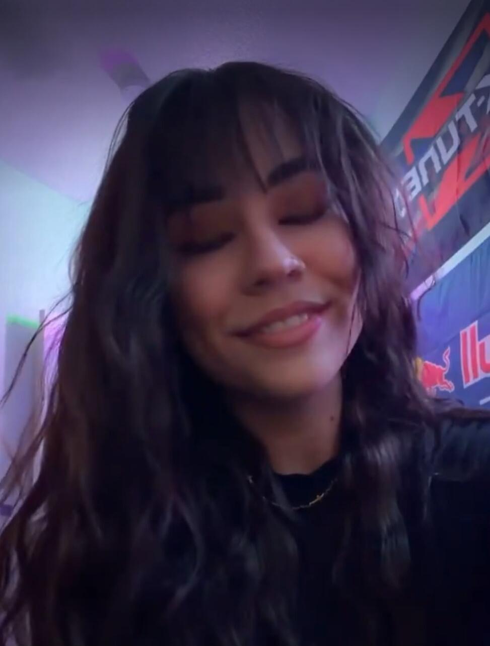A young woman with bangs smiles with her eyes closed. In the background, a Red Bull banner is visible.