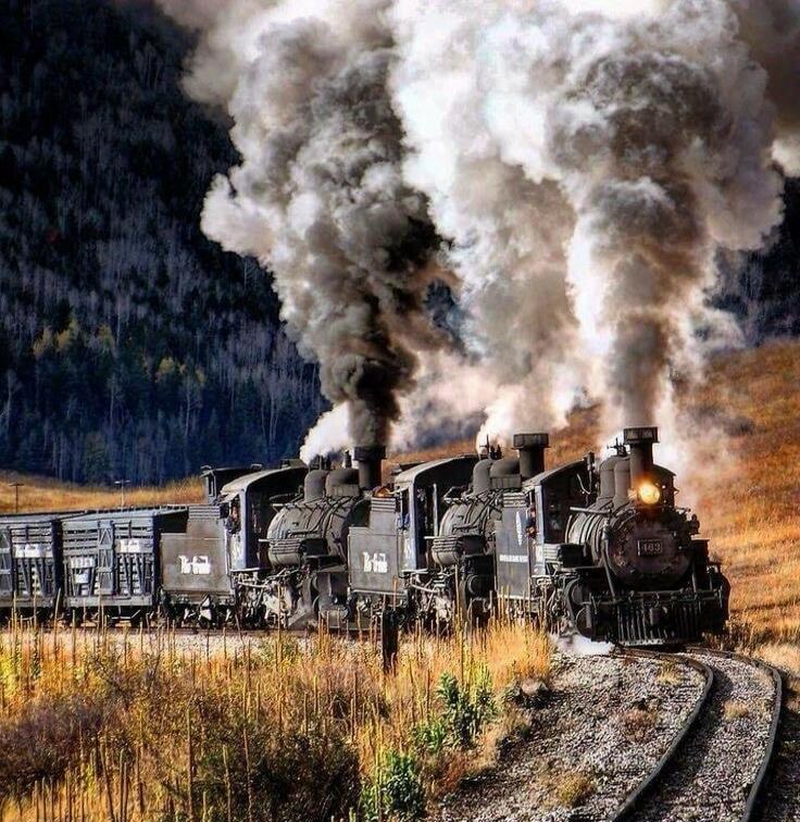 The image shows a train with multiple steam locomotives puffing out large clouds of smoke. The train is on a track that curves through a landscape with dry grass and trees in the background. The locomotives appear to be old-fashioned, with visible numbers on the front.