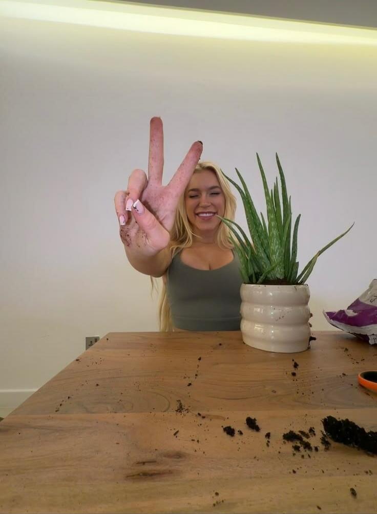 A blonde woman with a dirty hand making a peace sign, smiling, next to an aloe vera plant on a table with scattered soil.