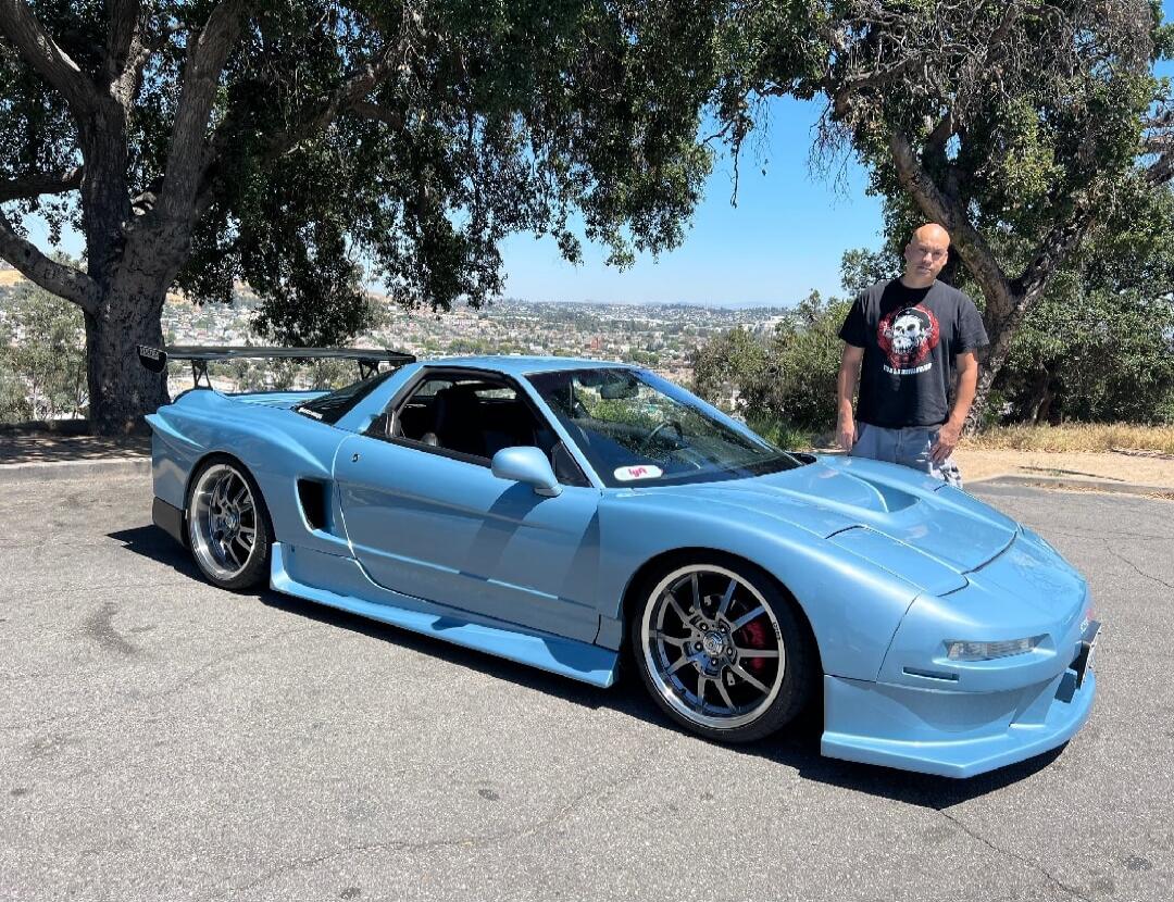 A blue sports car with a modified body kit parked on a paved area, a man standing beside it, with trees and a scenic hill in the background.