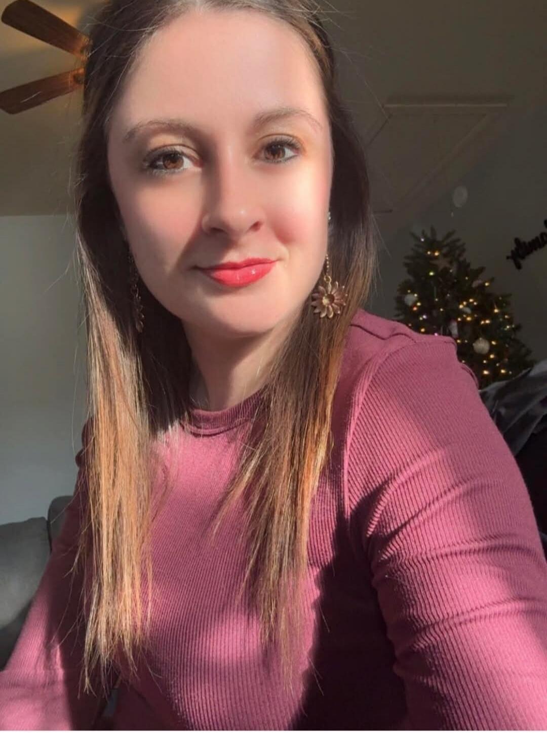 Portrait of a woman wearing a pink long-sleeve top, with a Christmas tree in the background.