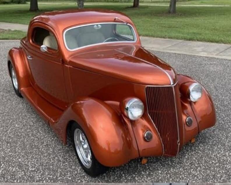 A classic, custom-painted orange-copper hot rod car is parked on an asphalt surface. The vehicle features a sleek, vintage design with prominent fenders, a vertical grille, and chrome accents. Green grass and trees are visible in the background under a bright sky.