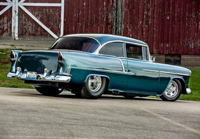 A vintage teal blue classic car, two-tone paint, parked on a road beside a red barn.