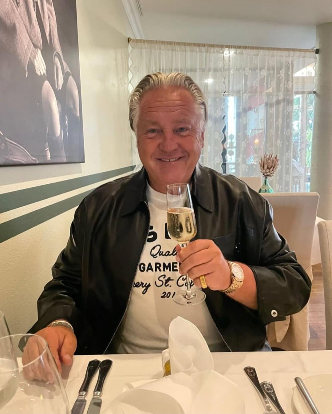 A smiling man in a black leather jacket and a white t-shirt is sitting at a restaurant table, holding a glass of champagne. The t-shirt has text: 'Quality GARMENT Luxury St. Cap 201'.