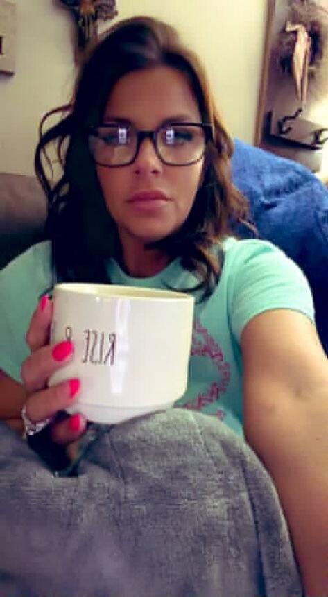 A woman wearing glasses and a light blue t-shirt holds a white mug with 'RISE' written on it. She has pink nail polish and a ring.