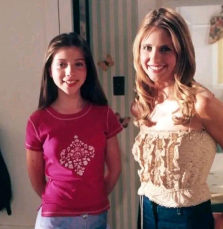 Two women, one younger and one older, smiling at the camera. The older woman has blonde hair and is wearing a light-colored, ruffled top. The younger woman has dark hair and is wearing a pink t-shirt with a white design.