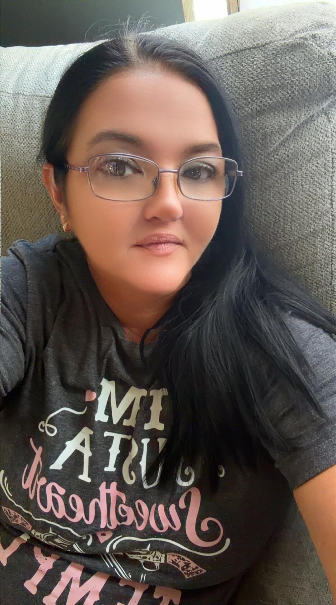 A woman is taking a selfie. She is wearing glasses and a dark t-shirt with partially visible, mirrored text that appears to say 'MI ATU' and some cursive text below, possibly 'Sweetheart' or 'Southern', along with images of two revolvers.