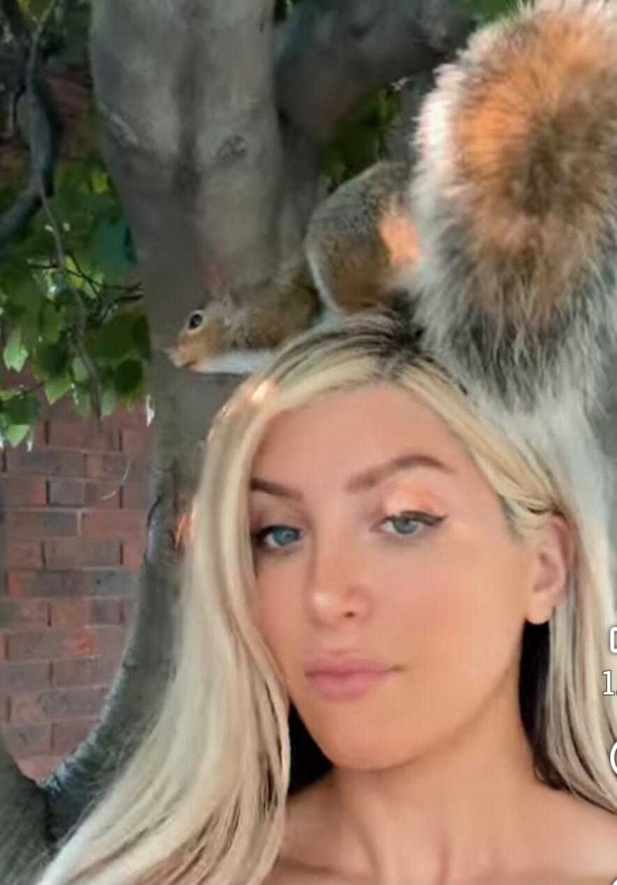 A woman with a squirrel perched on her head in front of a tree.