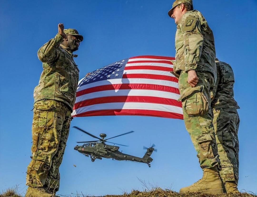 Two soldiers stand facing each other holding an American flag with a helicopter flying underneath.