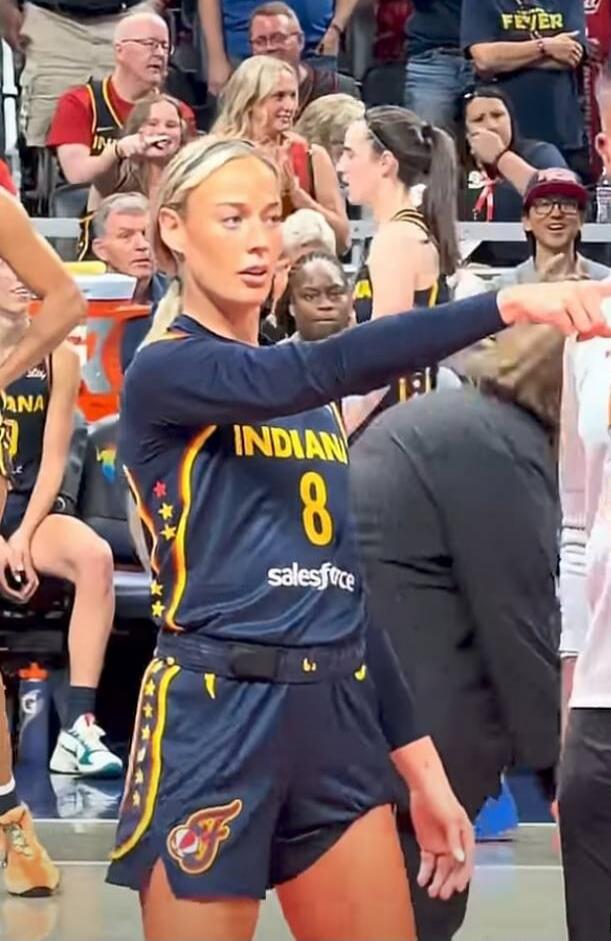 A female basketball player wearing a dark blue uniform with 'INDIAN' and '8' on it, along with 'salesforce', is pointing to the right during a game. Spectators are visible in the background. Other text visible includes 'FEVER' and 'ANA'.
