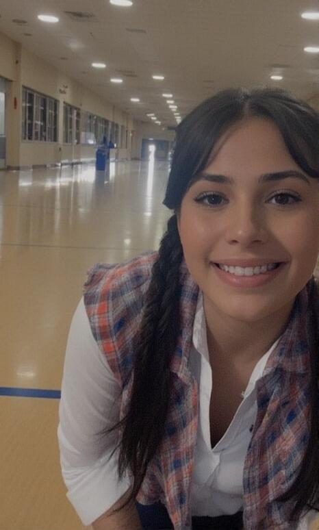 A smiling young woman with braided hair wearing a white shirt and a plaid sleeveless vest in a large indoor hallway.