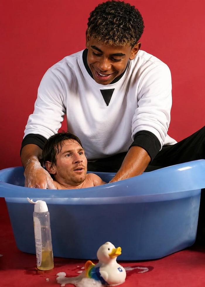 A man with the head of Lionel Messi is being bathed in a small blue tub by another smiling man. A rubber duck with rainbow wings is on the floor.
