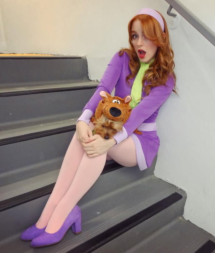 A young woman with red hair wearing a purple outfit sits on stairs, holding a fluffy plush dog.