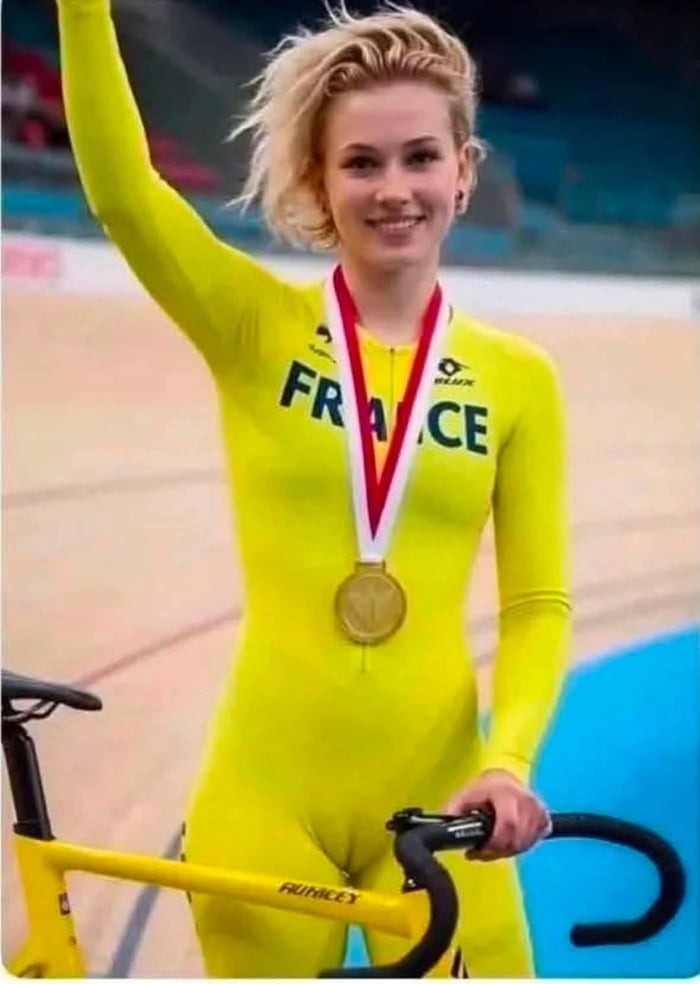 A female cyclist in a yellow full-body suit, wearing a gold medal, holding a bicycle, and smiling. The text 'FRANCE' is visible on her suit.