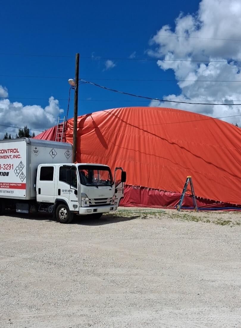 CONTROL PMENT, INC. 8-3291 TION REAL ESTATE INSPECTION SERVICES FREE ESTIMATES COL PEST CONTROL. A white truck from a pest control company is parked next to a large building covered with an orange fumigation tarp. A ladder is visible near a utility pole under a blue sky with clouds.