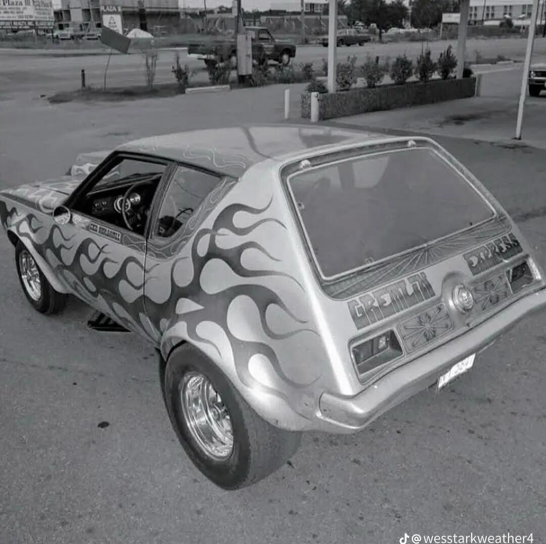 A silver hatchback car with large black flame graphics along the sides and rear, parked outdoors. The rear window and taillights are visible; wheels have polished rims.