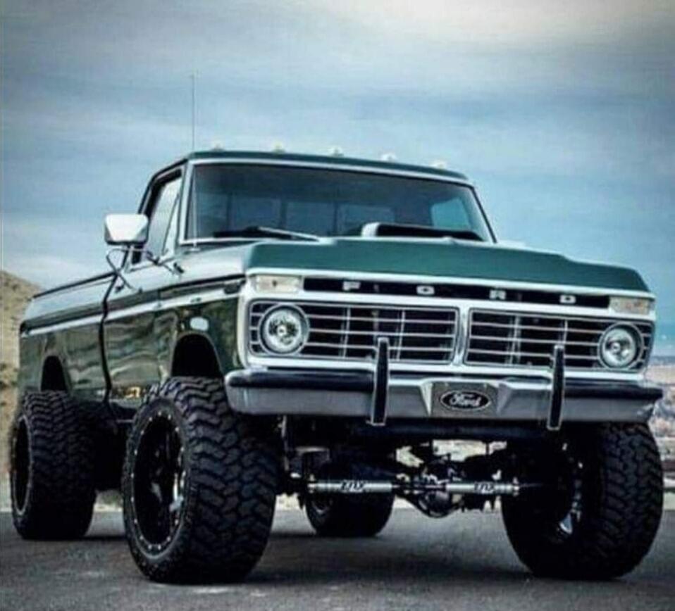 A green classic Ford pickup with oversized off-road tires and a lifted suspension, facing forward on a road with a cloudy sky.