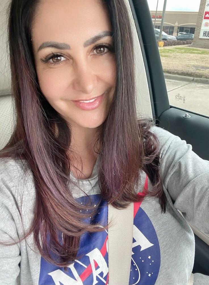 A woman in a car wearing a NASA sweatshirt. The NASA logo is visible on her shirt. In the background, a building with some text like 'HUB' is visible.