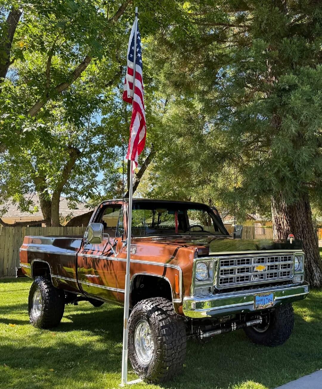 Orange vintage pickup truck with an American flag mounted on a pole in a grassy, tree-shaded yard.
