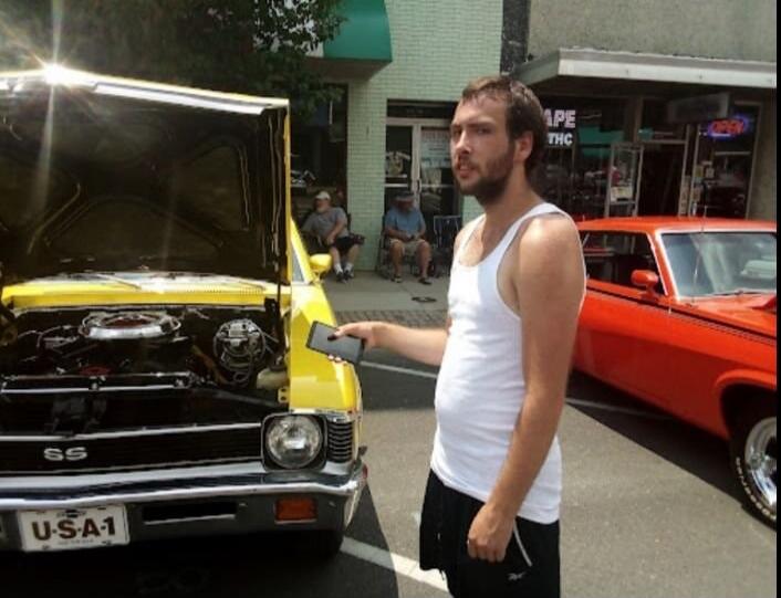 USA-1 license plate on yellow car; man in white tank top holding a phone next to a yellow vintage car with its hood open. Background shows a street with other cars and storefronts.