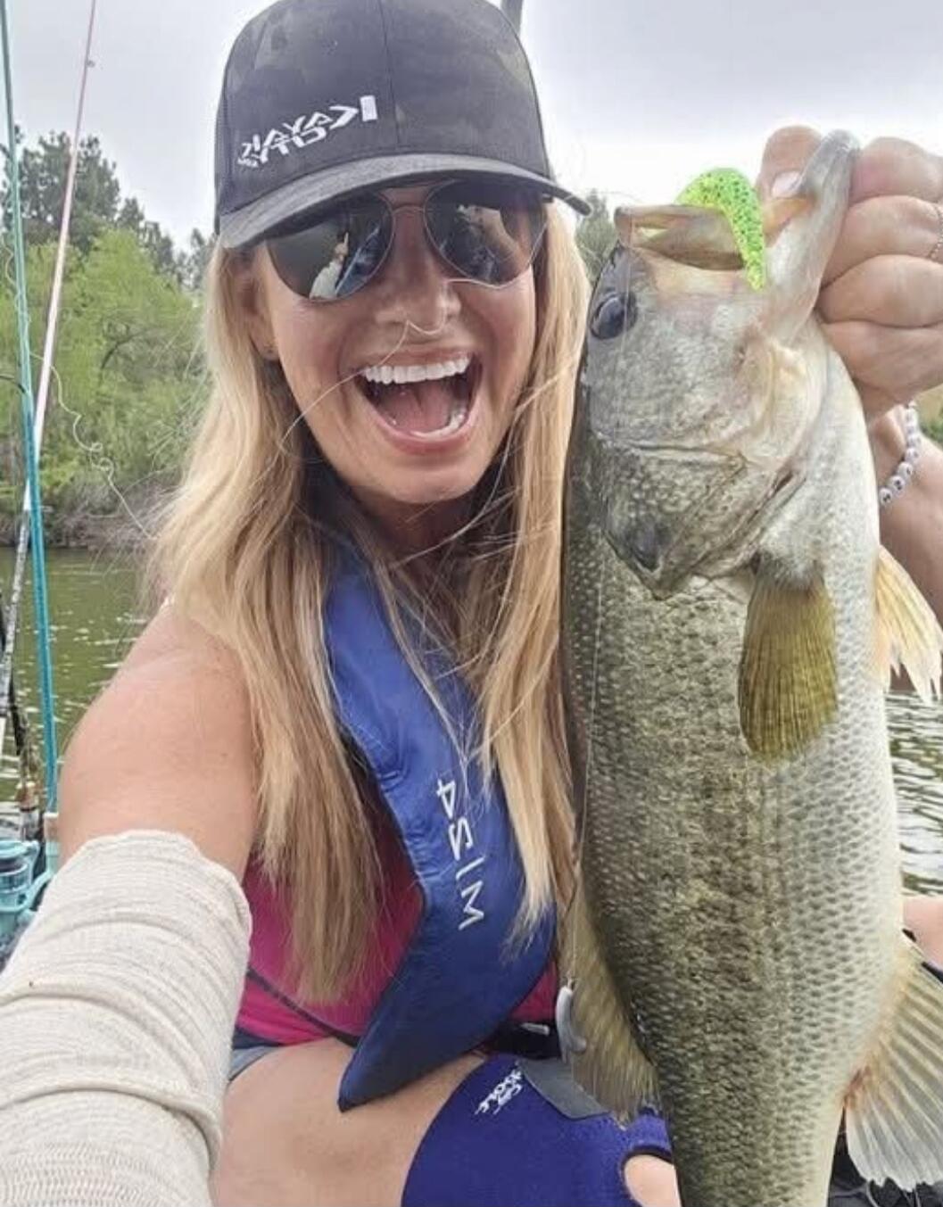 A woman in a life vest and a cap is holding a large bass fish with a green lure in its mouth. She is smiling widely, showing excitement. Fishing rods are visible in the background, suggesting she is on a boat or kayak on a body of water. The cap has text that appears to be 'KAYAK' or similar, and the life vest has 'PSIM' written upside down.