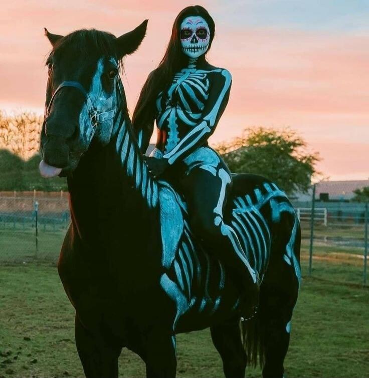 A woman with skeleton body paint and sugar skull face paint is riding a black horse, which also has skeleton paint. The horse has its tongue slightly out. The scene is set against a sunset sky in a field.