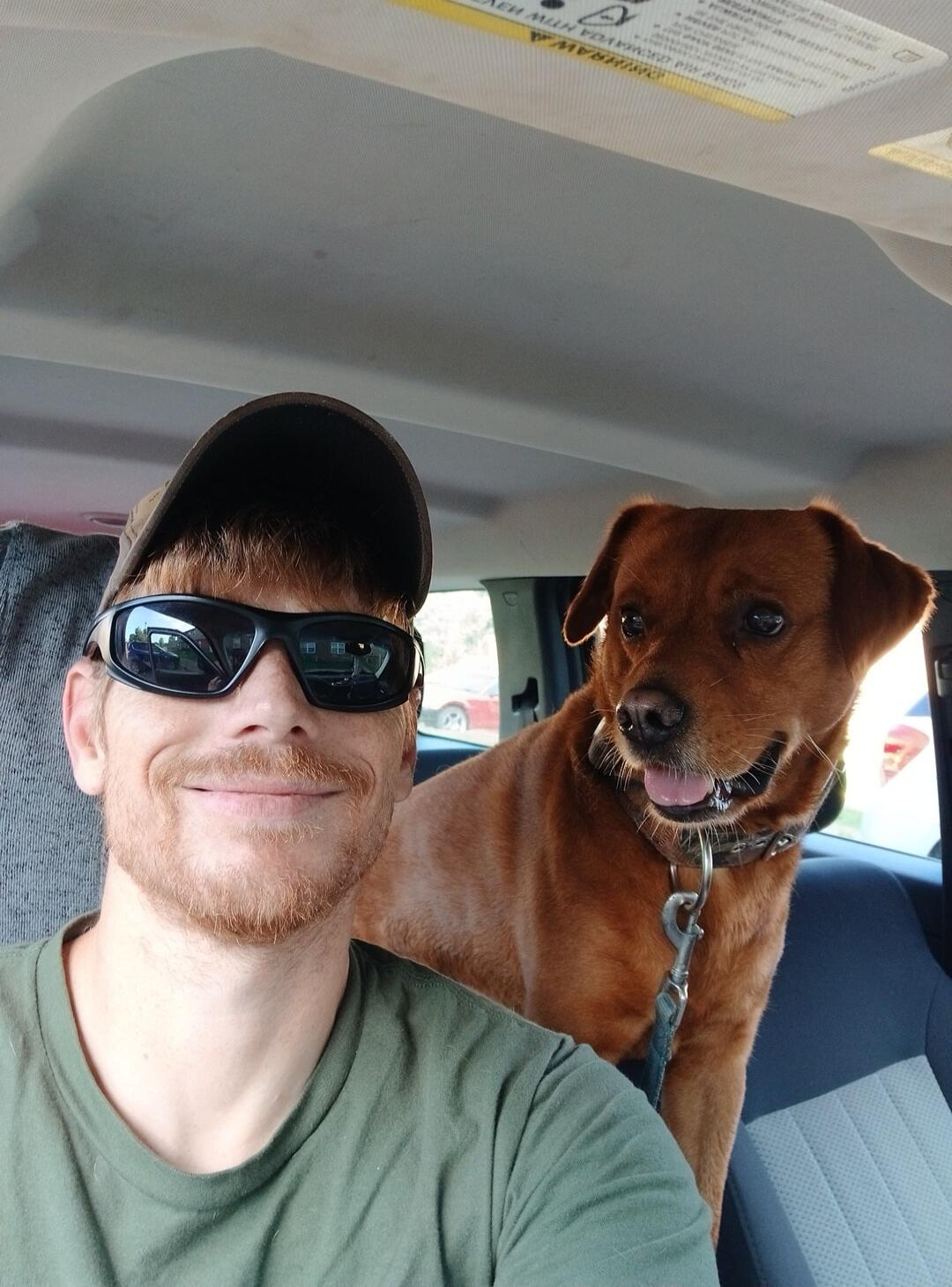 A man and a dog are in a car. The man is taking a selfie, smiling, wearing a cap and sunglasses. The dog is reddish-brown, sitting next to him, looking happy with its tongue slightly out. A warning label is visible on the car's sun visor, but the text is unreadable.