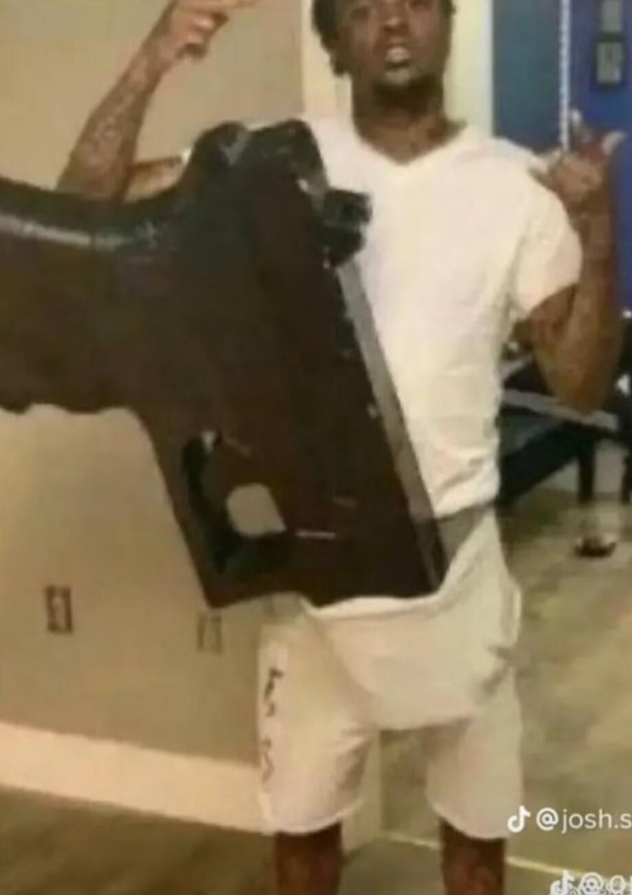 The man is holding a large, black object that resembles a gun. He is wearing a white t-shirt and white shorts. He is pointing his finger upwards with his left hand and making a hand gesture with his right hand.