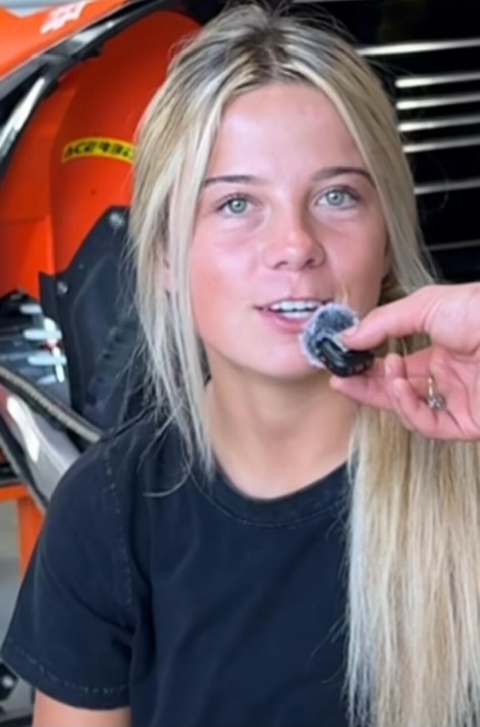 A young blonde woman is being interviewed. A microphone is held near her mouth. The brand 'ACERBIS' is visible on an orange object in the background.