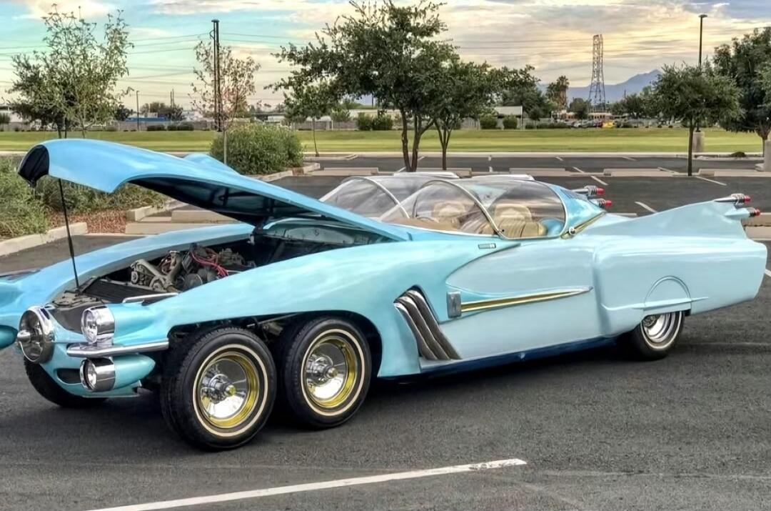 A light blue vintage car with an open hood and a custom body. The car has a unique tri-axle rear setup with two rear wheels on each side, giving it six wheels in total. It features a chrome grille, chrome accents, and a bubble canopy-style dual-seat cockpit. The car is parked in an empty lot with trees in the background.