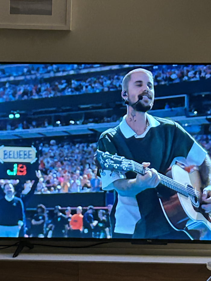 A man is performing live music, singing and playing an acoustic guitar on stage in front of a large crowd. A sign in the background reads 'BELIEBE JB'.