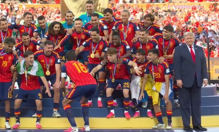 A group of soccer players in red and yellow uniforms, likely the Spanish national team, are celebrating on a podium with medals. Donald Trump is standing next to them.