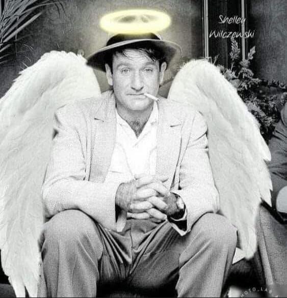 A man in a light suit sits with his hands clasped, wearing a hat with a halo above and angel wings behind him; he is smoking a cigarette.