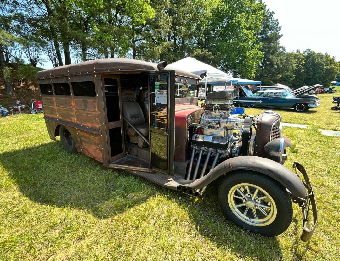 Rusty vintage hot rod-style vehicle with an exposed engine and cabin doors open, parked on a grassy field at a car show.