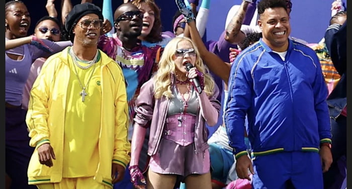 A group of people, including Madonna, Ronaldinho, and Ronaldo, are seen together. Madonna is in the center, holding a microphone and singing, dressed in a pink and purple outfit with a corset and shorts. To her left, Ronaldinho is smiling, wearing a yellow tracksuit and sunglasses. To her right, Ronaldo is also smiling, wearing a blue tracksuit. Th