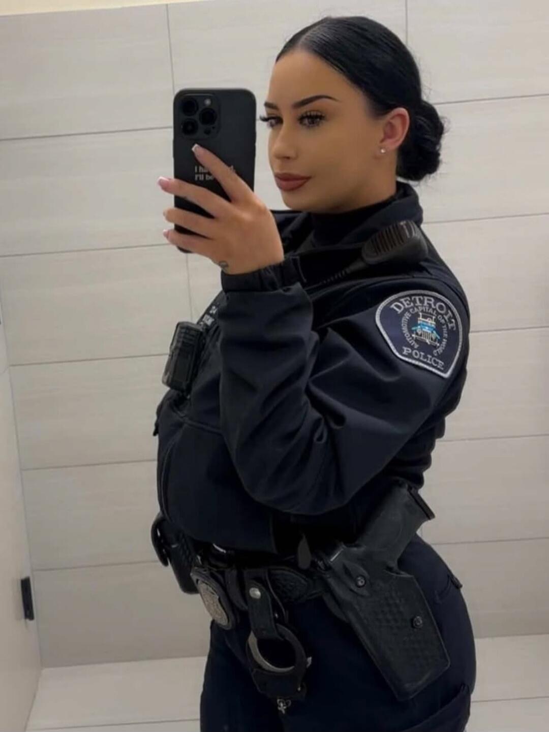 A woman in a Detroit Police uniform takes a mirror selfie. She is wearing a dark uniform jacket over a turtleneck, with a duty belt holding a handgun in a holster and handcuffs. The text on her phone case reads 'I hate I'll be'.