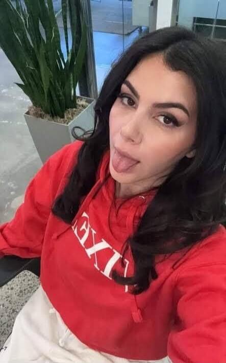 A woman in a red hoodie with white text 'EXI' visible, sticking out her tongue.