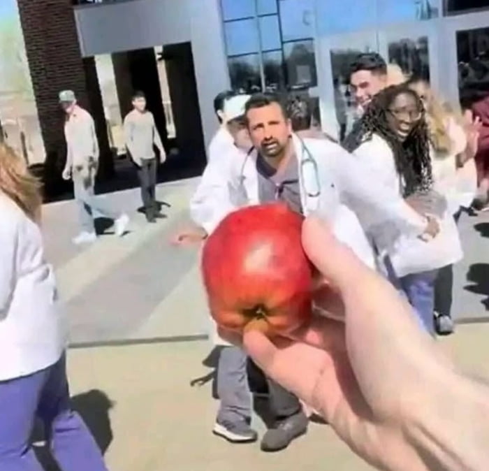 An apple is held in the foreground, with a group of people in white coats (likely medical staff) outside a building in the background.